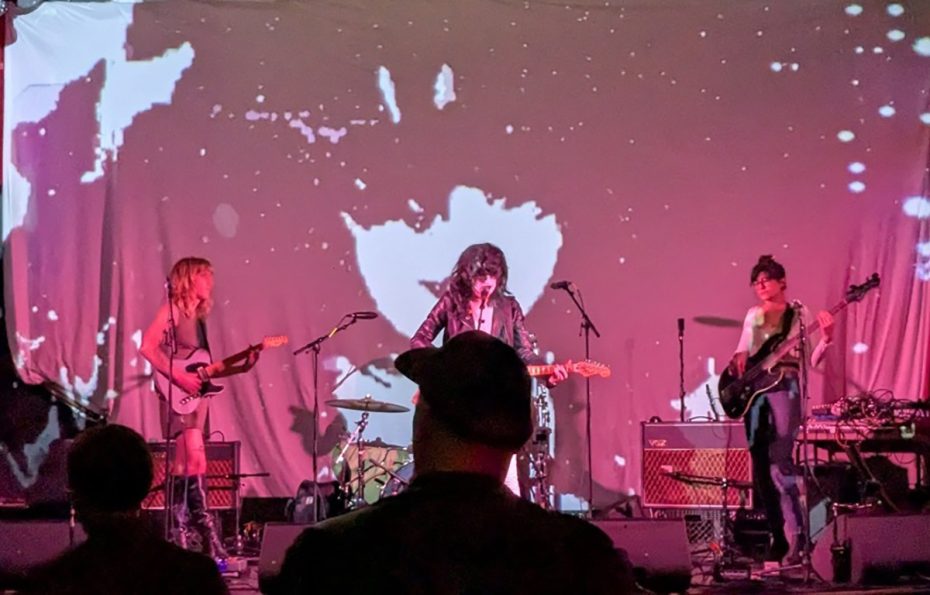 A band performs on stage against a backdrop of abstract visuals. Three musicians play electric guitars and sing, with an audience watching in front.