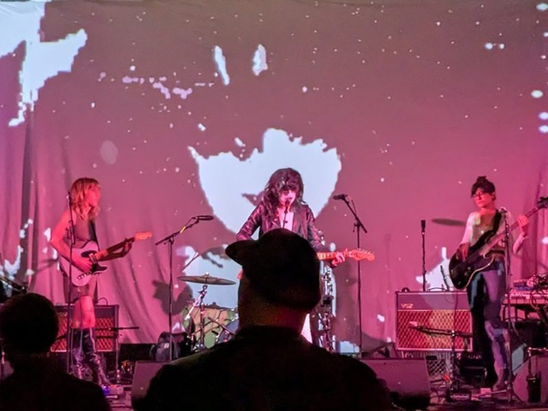 A band performs on stage with guitars and drums. A colorful background projection enhances the scene. Audience members watch in the foreground.