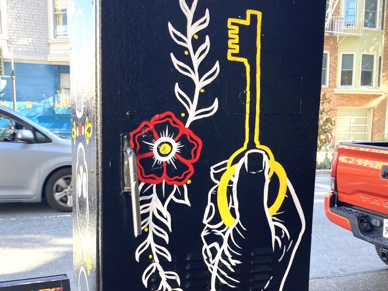 Street art on a utility box shows a hand holding a yellow key, surrounded by a vine with red flowers against a black background.