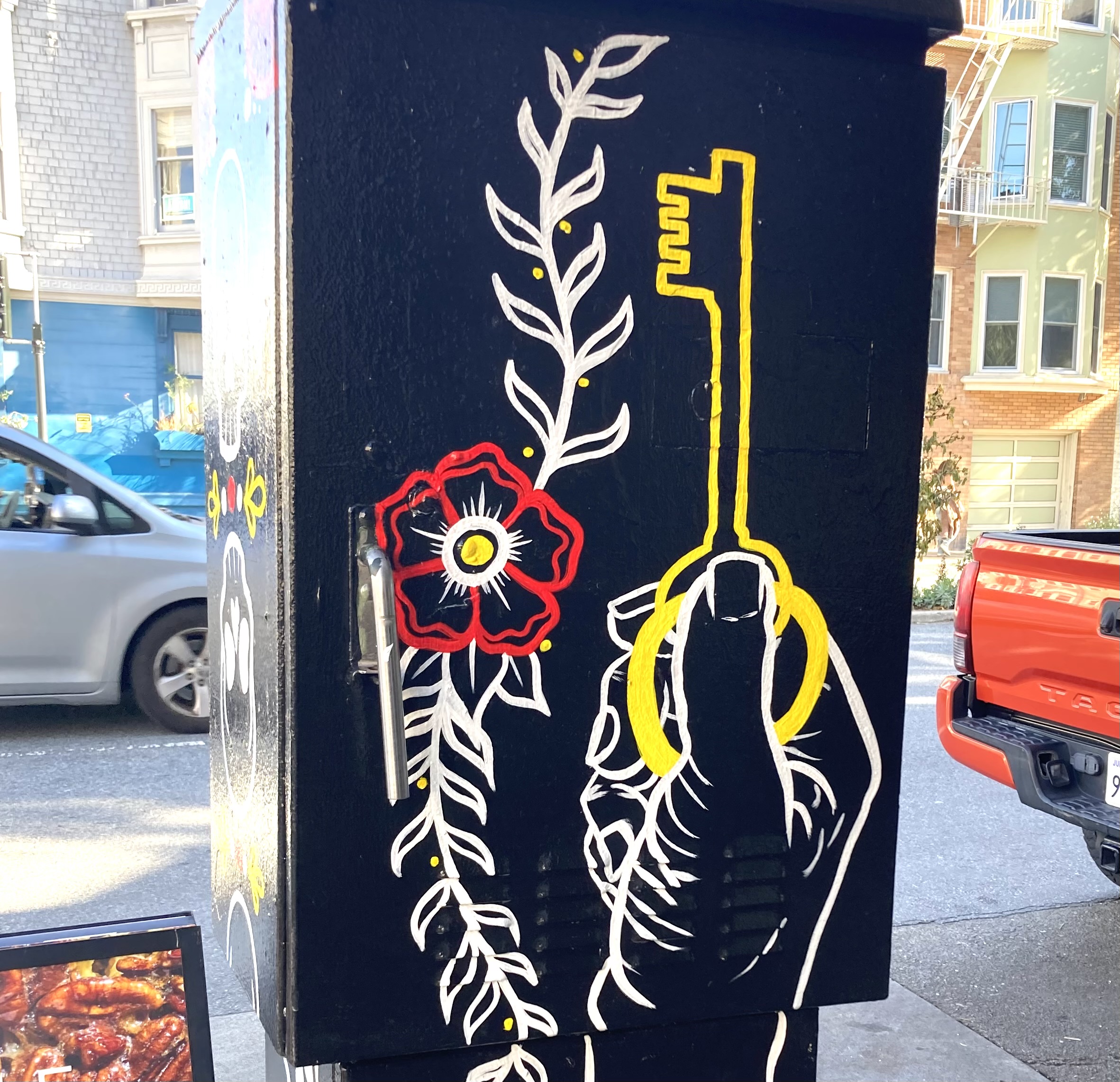 Street art on a utility box shows a hand holding a yellow key, surrounded by a vine with red flowers against a black background.