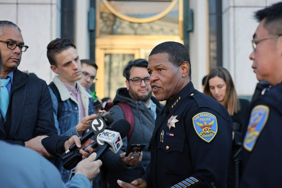 A police officer in uniform, emphasizing the importance of mental health awareness, addresses a group of journalists holding microphones and taking notes outside a building.