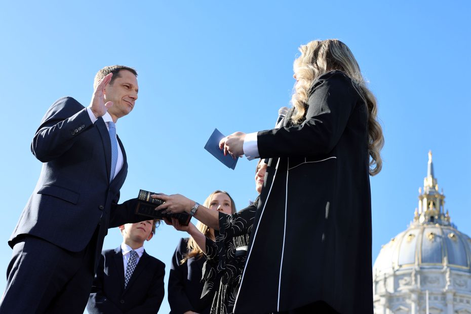 A person in a suit is being sworn in outdoors by another person holding a book, with a group of people standing nearby and a domed building in the background.