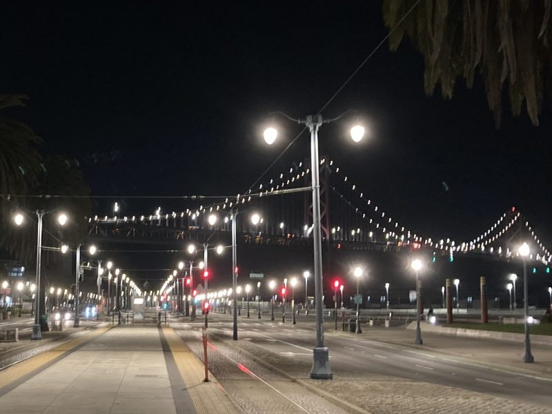 Night view of a lit-up suspension bridge with lights reflecting on the water. Empty street and tram tracks in the foreground.