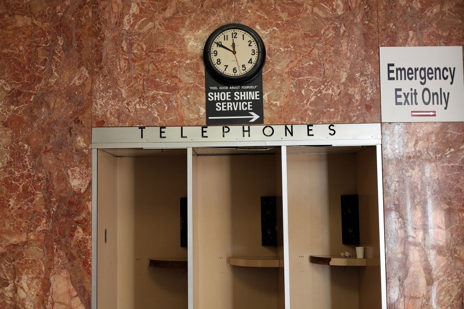 Empty telephone booths with a clock above and signs for a shoe shine service and emergency exit on a marble wall.