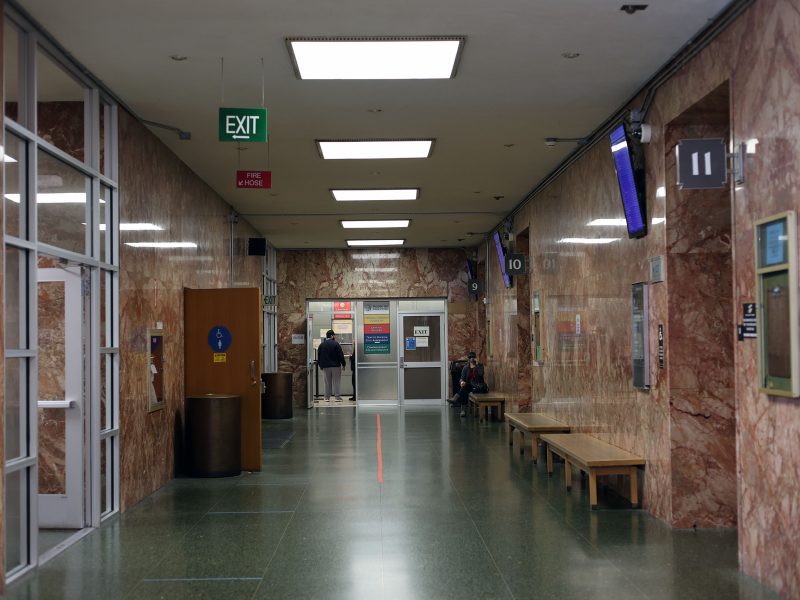 A hallway with marble walls, green floors, exit signs, and benches. People are seated and a person stands by an information board near the doorway at the end.