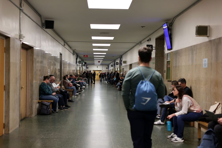A long hallway with people sitting on benches along the walls. One person is walking down the center. The hallway has various doors and overhead signs.