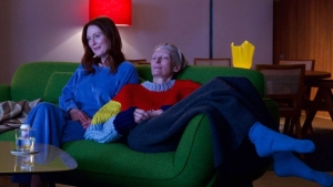 Two people relax on a green sofa in a cozy room with a red lamp. One wears blue, the other a red sweater. A drink and a yellow item are nearby.
