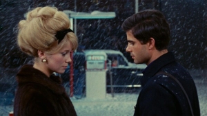 A man and a woman with a beehive hairstyle stand facing each other outdoors in the snow, next to a vintage car and a gas pump.