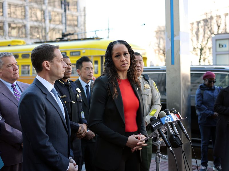 People standing at a press conference with microphones, including a woman in a red shirt speaking. A yellow vehicle is in the background.