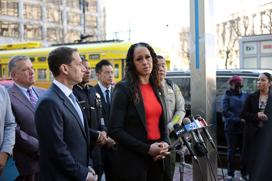 People standing at a press conference with microphones, including a woman in a red shirt speaking. A yellow vehicle is in the background.