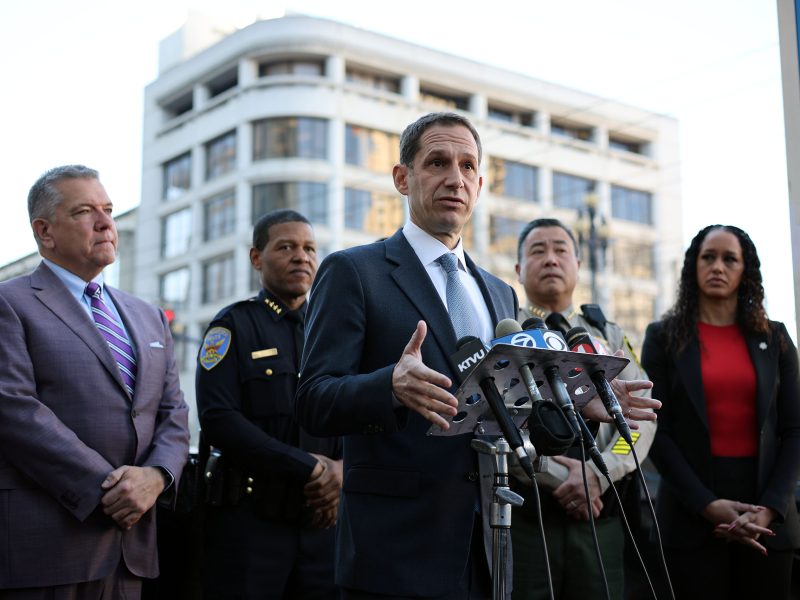 A man in a suit speaks at a podium with microphones, flanked by four individuals. A city building is in the background.