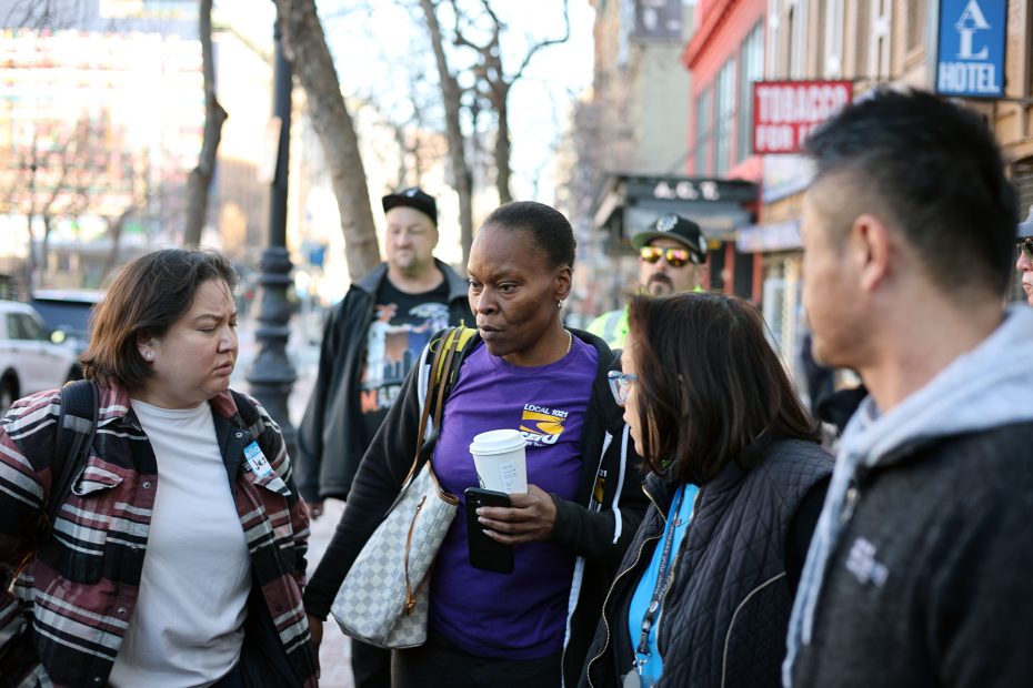 A group of people stands on a sidewalk, engaging in conversation. Some hold coffee cups and wear casual clothes, with buildings and trees visible in the background.