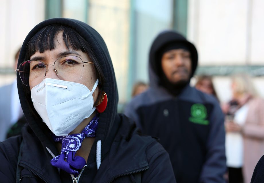 A person wearing a face mask and hood stands in the foreground of a crowd, with another person wearing a hood visible in the background.