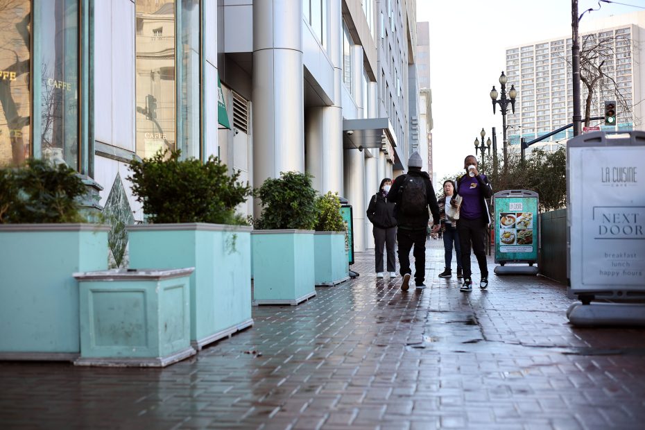 People walking on a wet city sidewalk lined with large planters and buildings.