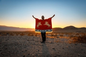 Person standing in a desert landscape at sunset, holding a red blanket with horse designs. Arms are outstretched.