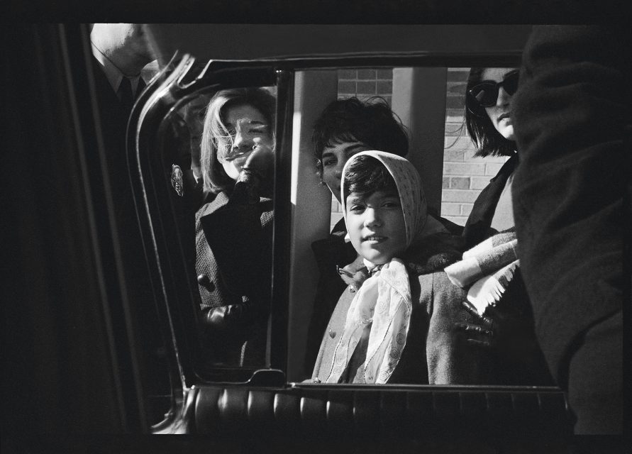 A group of people, including a child wearing a headscarf, is seen through a car window, surrounded by adults in formal attire, as if they are heading to one of the city's prestigious museums.