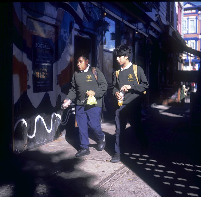 Two students in uniforms walk along a street with snack bags and drinks. A mural is visible on the building behind them.