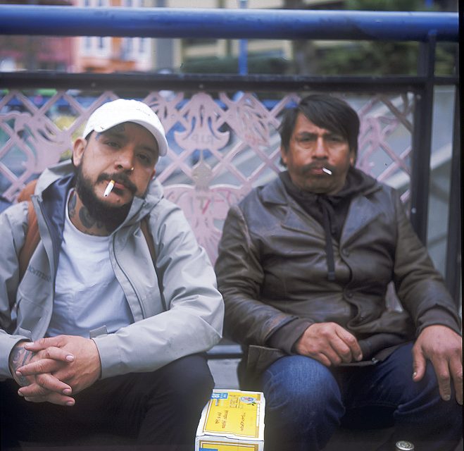 Two men sitting on a bench, both with cigarettes in their mouths; one wears a cap and jacket, the other wears a leather jacket. A box with text is on the bench in front of them.
