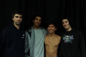 Four people standing together against a dark background; one is shirtless while the others are wearing casual clothing.