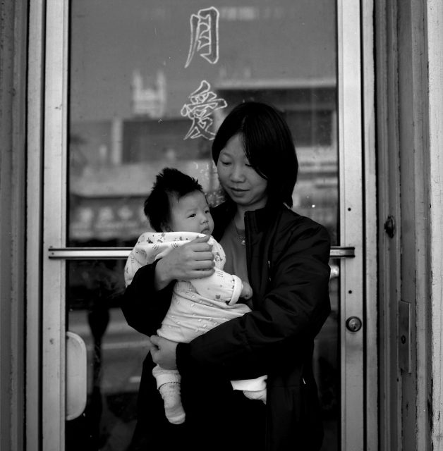 A woman stands in front of a glass door, holding a baby wrapped in a patterned blanket. Chinese characters are visible on the door.