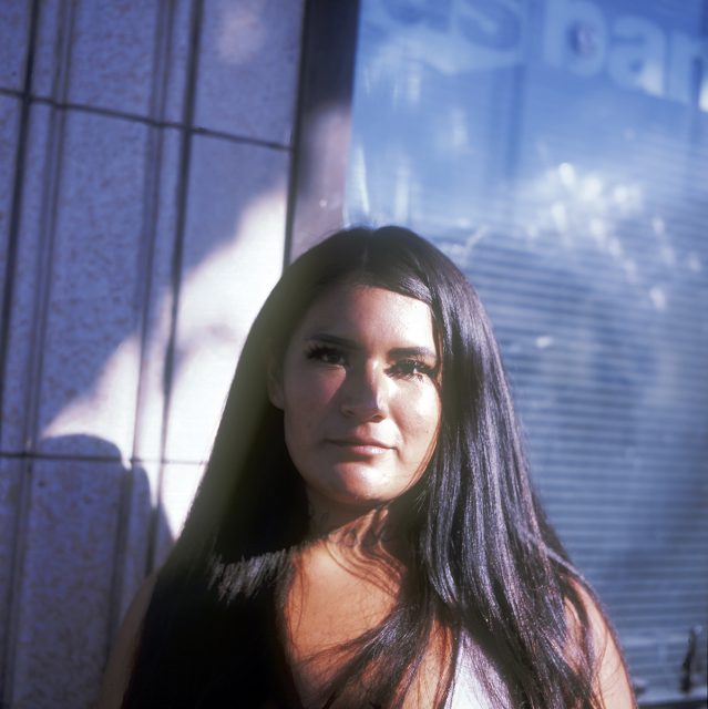 A woman with long dark hair stands outside a building, partially in sunlight, against a glass window with reflections.
