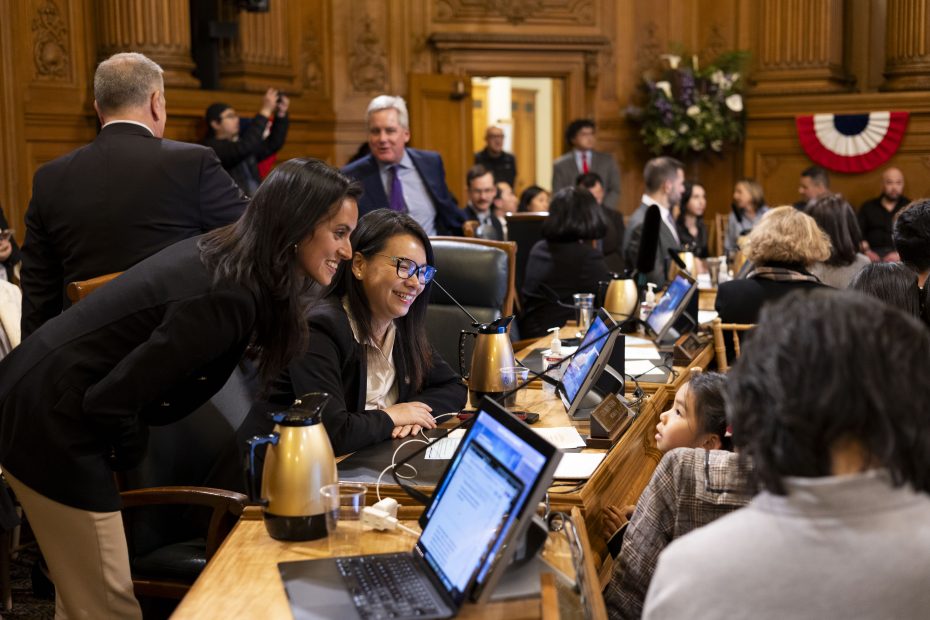 A group of people in a meeting room with laptops and documents on desks. Two women are smiling and conversing, while others are seated or standing in the background.