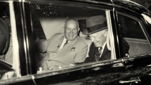 Two men in suits sit smiling in the back seat of a car. One wears a hat, and they appear to be engaged in conversation.