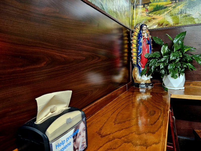 A tissue dispenser and a plant with a Virgin Mary statue are placed on a wooden counter against a mural wall.