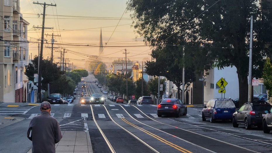 City street at sunset with cars, power lines, and trees. A person stands in the foreground, and a distant hill is visible under a colorful sky.