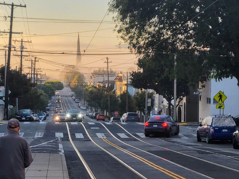 City street with cars, power lines, and a person walking on the sidewalk in the foreground. Trees and buildings line the road, and a distant landmark is visible in the background.