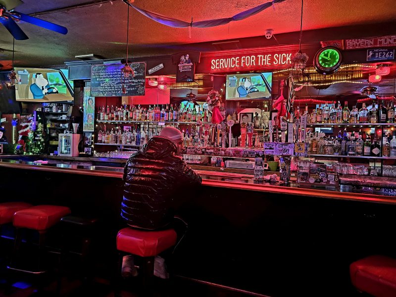 A person in a dark jacket sits alone at a dimly lit bar with a neon sign reading "Service for the Sick" above. The bar is cluttered, and TV screens show animated content.