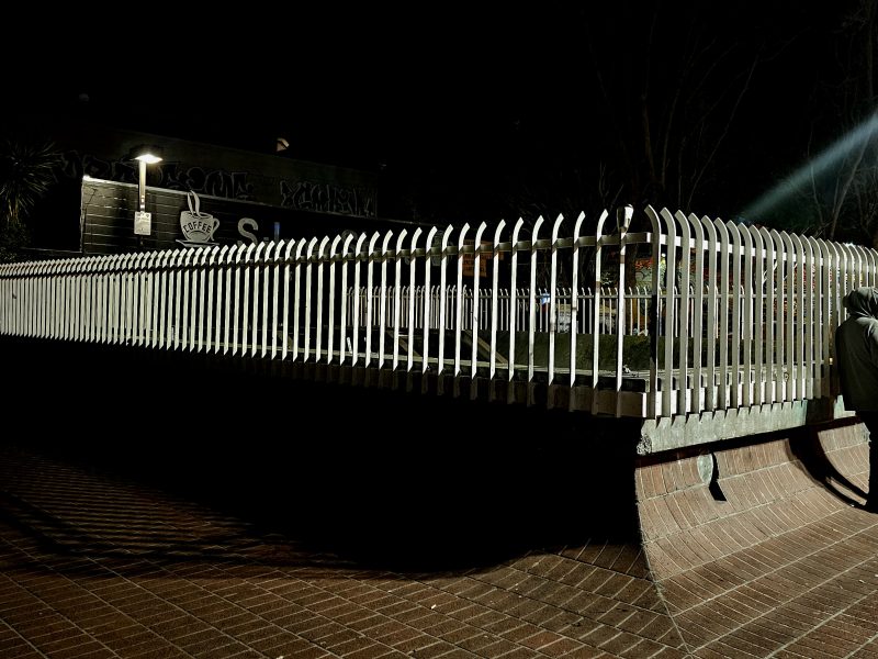 A person in a hoodie stands beside a fenced structure at night, with another person sitting nearby on a brick-paved surface.