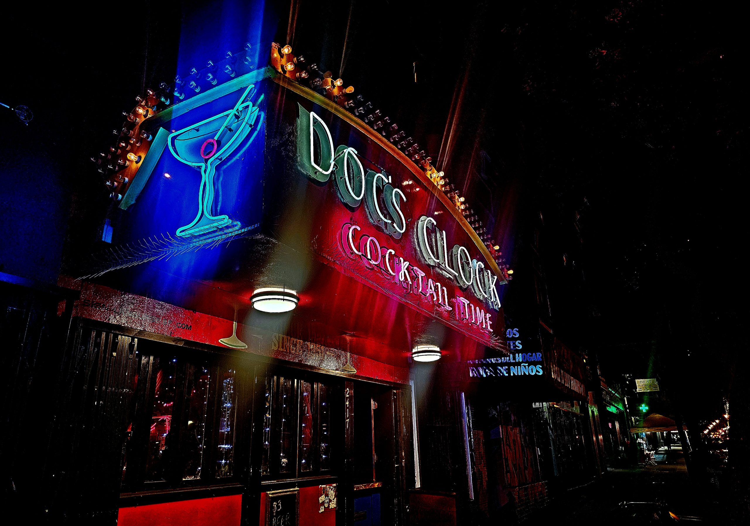 Neon sign for "Doc's Clock Cocktail Time" above a bar entrance, featuring a martini glass with an olive.