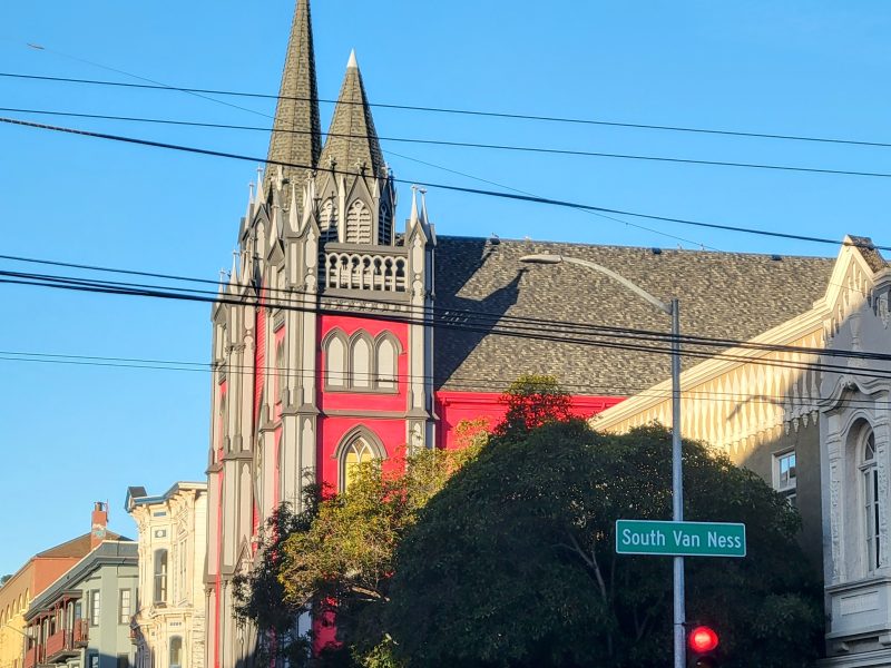 Gothic-style building with red and grey facade, featuring tall spires, located at an intersection with a street sign for South Van Ness, against a clear blue sky.