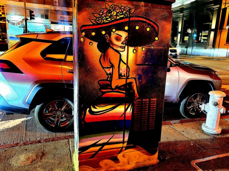 Colorful mural of a woman in a hat painted on a utility box at a city street corner, with cars and buildings in the background, at night.