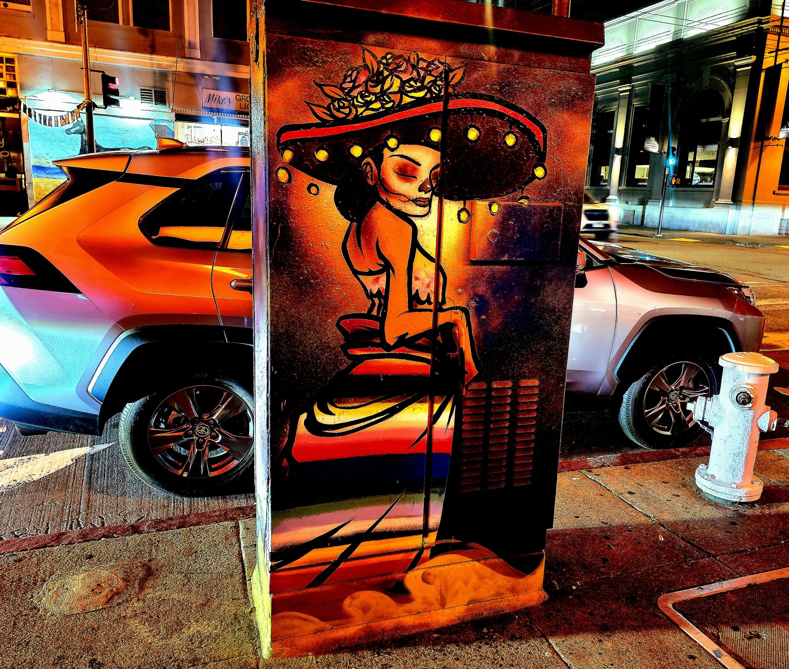 Colorful mural of a woman in a hat painted on a utility box at a city street corner, with cars and buildings in the background, at night.