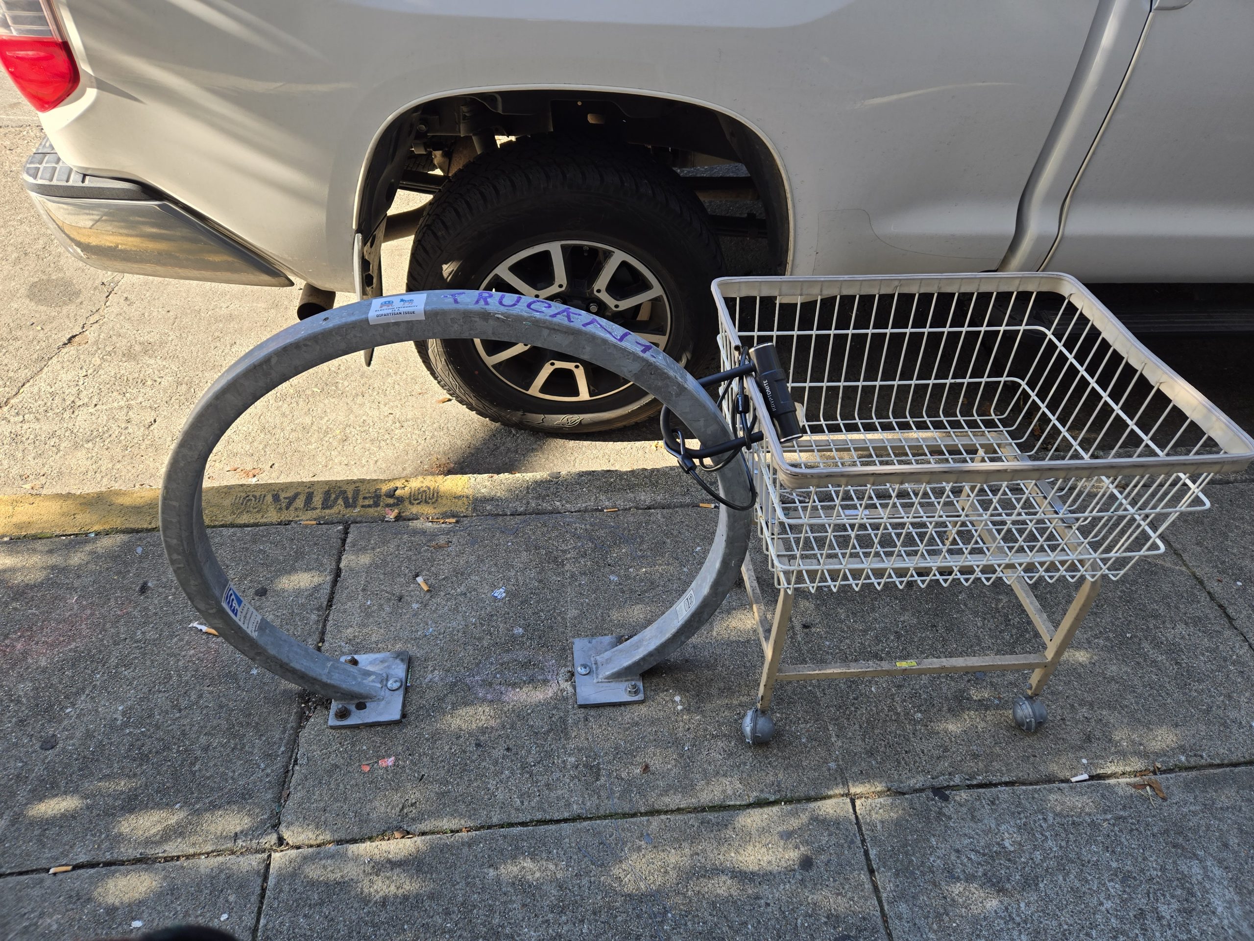 A shopping cart is chained to a circular metal bike rack on a sidewalk, next to a parked vehicle.