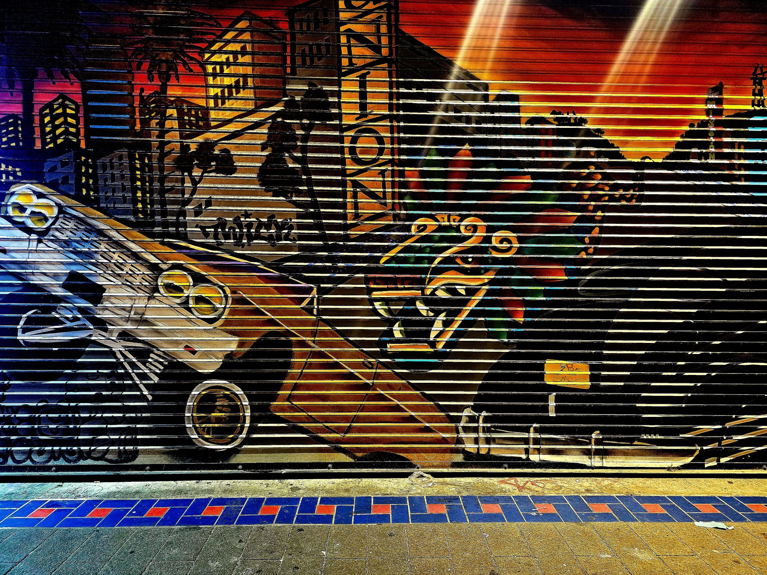 Colorful street mural depicting a cityscape with classic cars, palm trees, and a serpent motif, painted on a corrugated metal shutter.