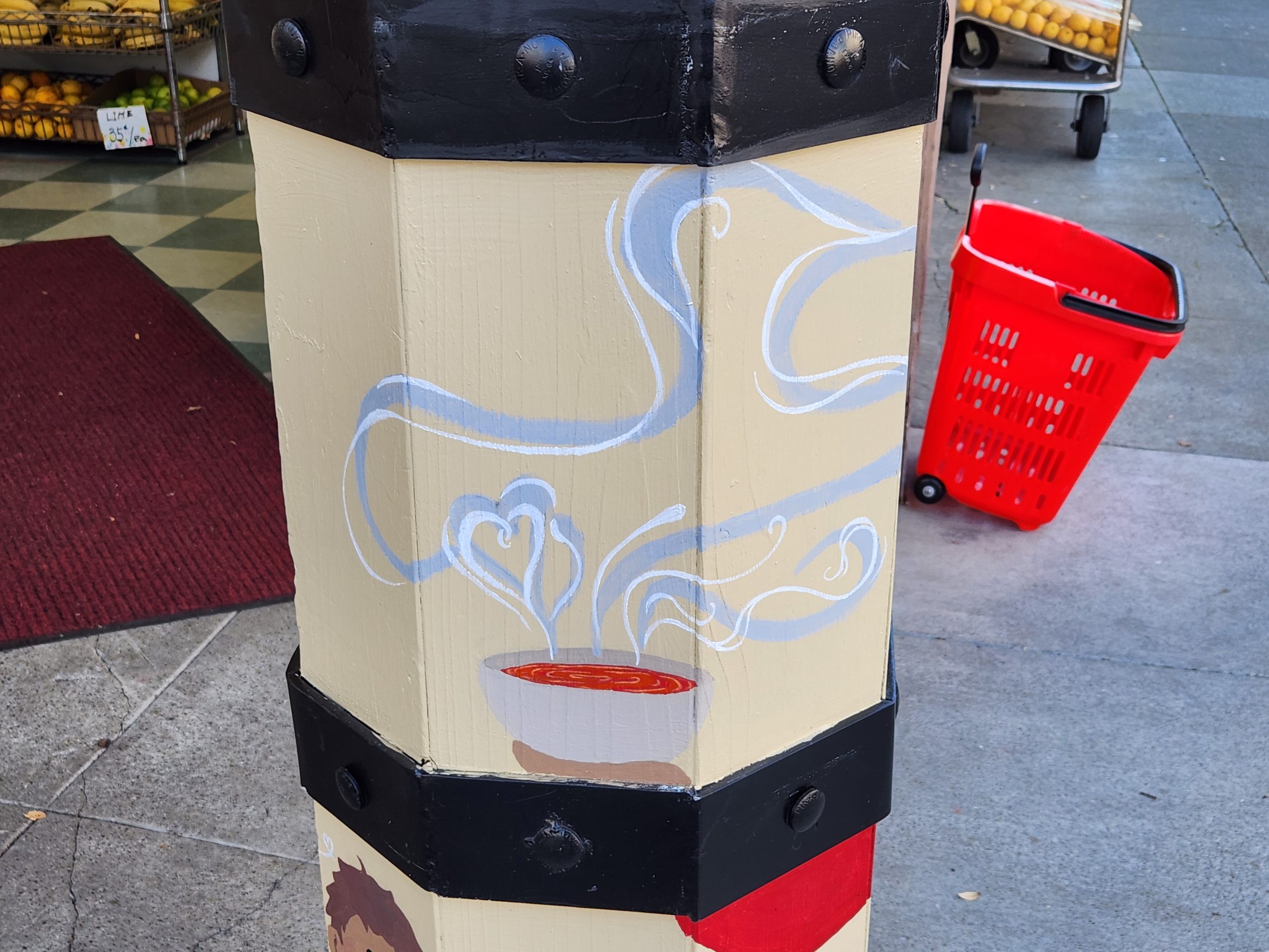 Illustrated utility pole with a steaming bowl of noodles design, near a red shopping basket on a sidewalk.