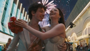 A couple joyfully embraces under fireworks, with bright lights and buildings in the background. The man holds flowers, and they both appear happy.