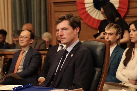 A group of people in business attire sit attentively in a government meeting room, with a large, patriotic banner in the background.