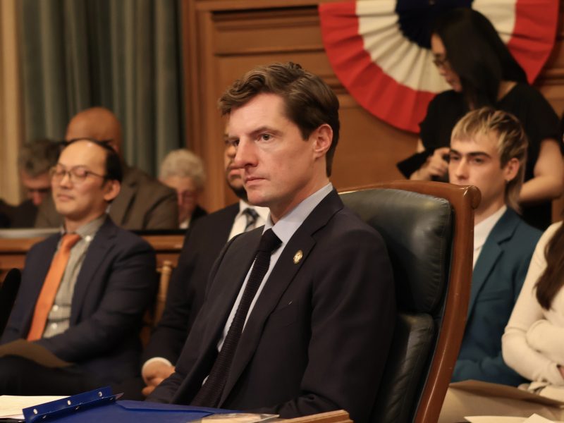 A group of people in business attire sit attentively in a government meeting room, with a large, patriotic banner in the background.