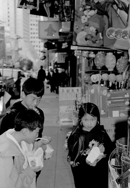 Three children stand on a busy city street, looking at items displayed outside a shop. The scene is in black and white.