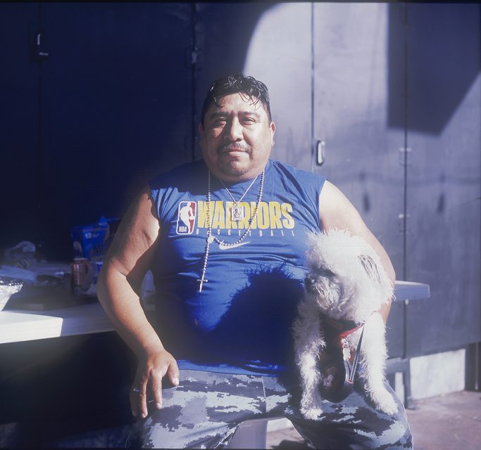 Man in a blue Warriors shirt sits on a bench with a small white dog on his lap.