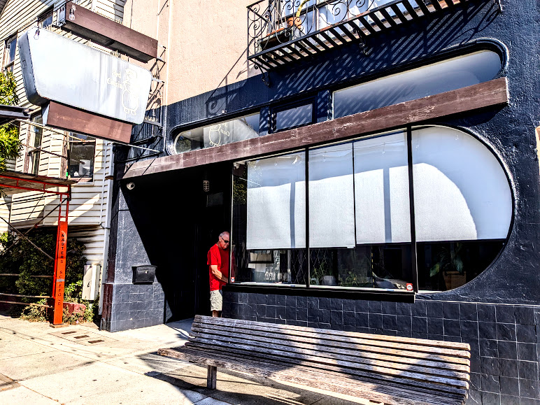 A person stands in front of a building with rounded windows and a bench on a sunny day.
