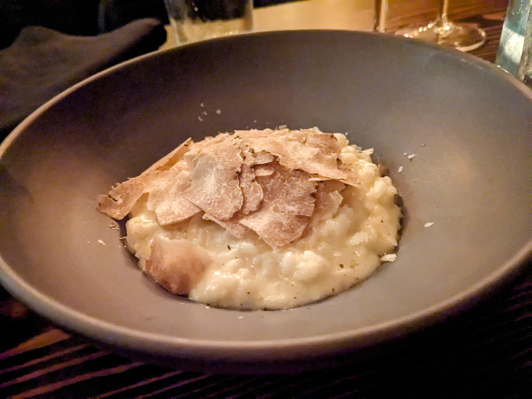 A bowl of creamy risotto topped with thin slices of truffle in a dark dish.