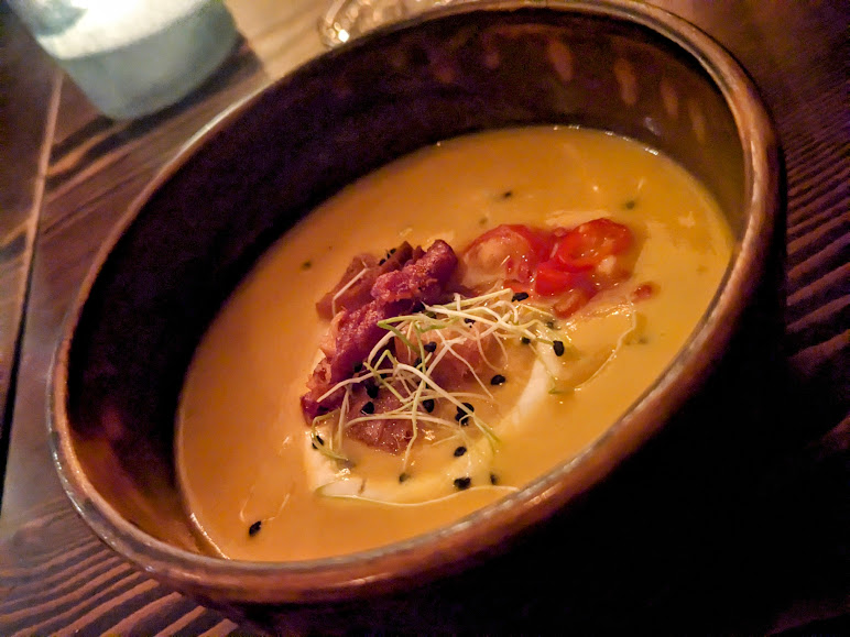 A bowl of creamy soup topped with crispy bacon bits, chopped vegetables, and sprouts on a wooden table.