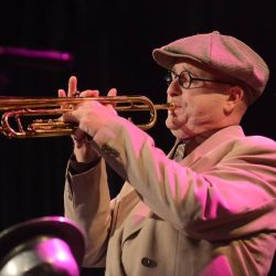A person in a beige suit and cap plays a trumpet on stage under pink lighting.