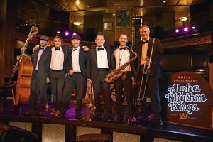 Six musicians in tuxedos with various instruments pose on stage with an "Alpha Rhythm Kings" sign in a dimly lit venue.