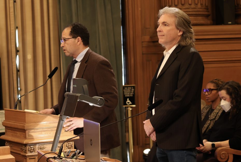 Two men stand in a formal setting, one speaking at a lectern with microphones, the other standing beside him in a blazer. Two seated individuals are visible in the background.